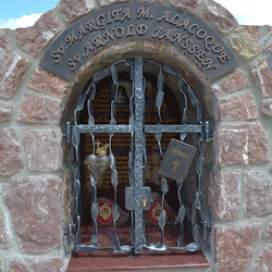 Kovaný pamätník. Sv. Margita M. Alacoque - srdce s plameňom, krížom a tŕňovou korunou, Sv. Arnold Janssen - Biblia